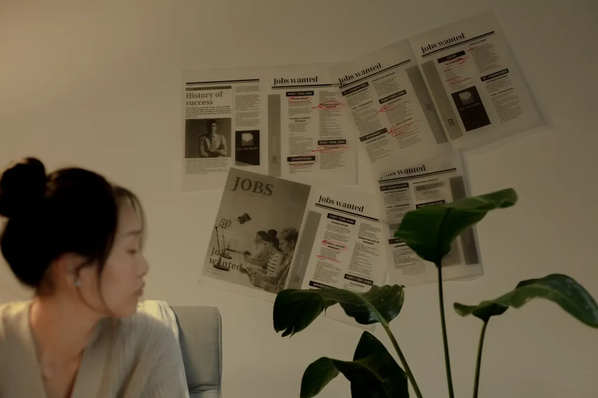 A woman sits pensively indoors beside a plant and job postings.
